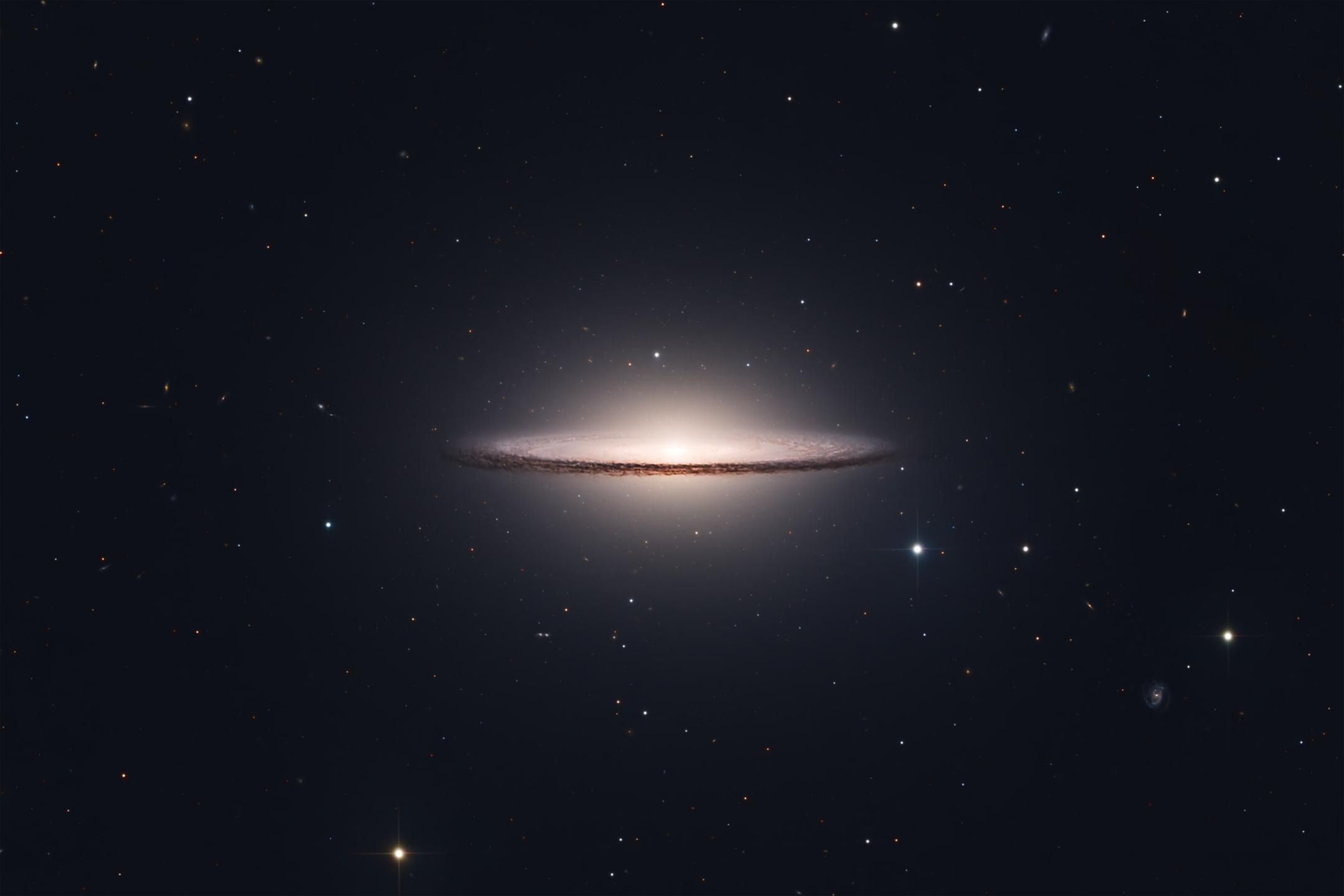 A galaxy seen almost edge-on: a brilliant white bulge crossed by a fine, sharply defined dark dust lane along its rim, its faint outer halo fading into an otherwise near-empty star field.