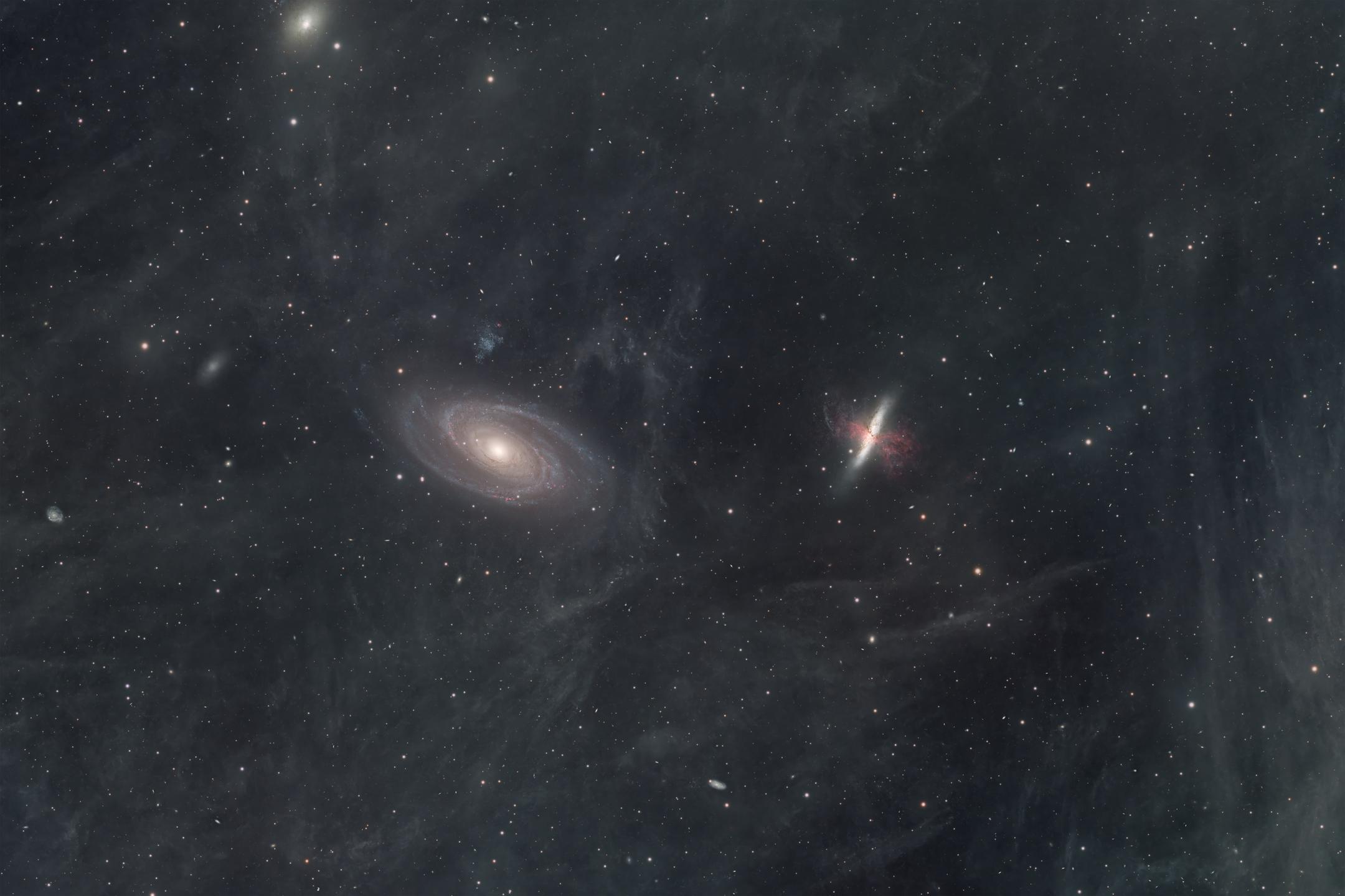Two galaxies against a field veiled in faint brown-grey dust — a broad face-on spiral on the left, and to its right a smaller galaxy seen edge-on and tilted, with red filaments erupting from either side of its centre.