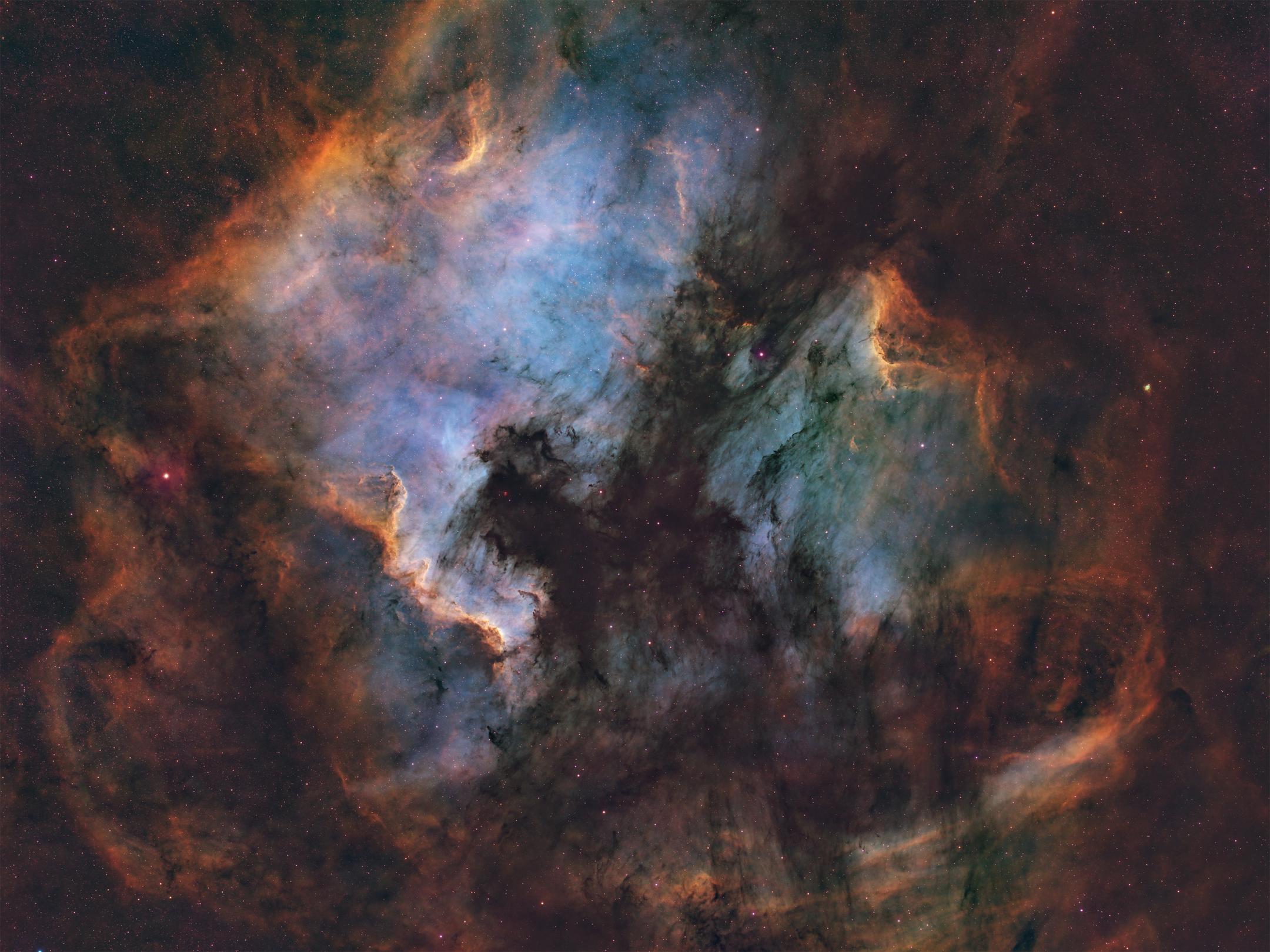 Two large emission clouds in rust-orange and blue-grey, their facing edges lit into bright ridges, separated by a broad dark dust rift whose outline curves down the centre of the frame like a coastline.